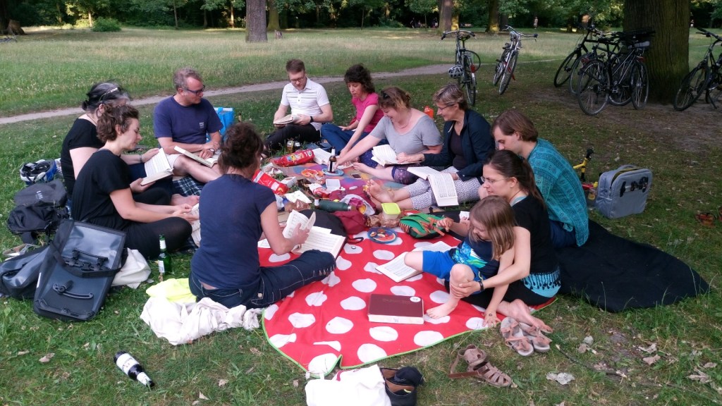 Eleven people sitting around a red and white-spotted rug in a Berlin park, with Sacred Harp song books on their knees, and food and drink visible on the rug. What may be one of Jan's uncomfortable shoes in the foreground.