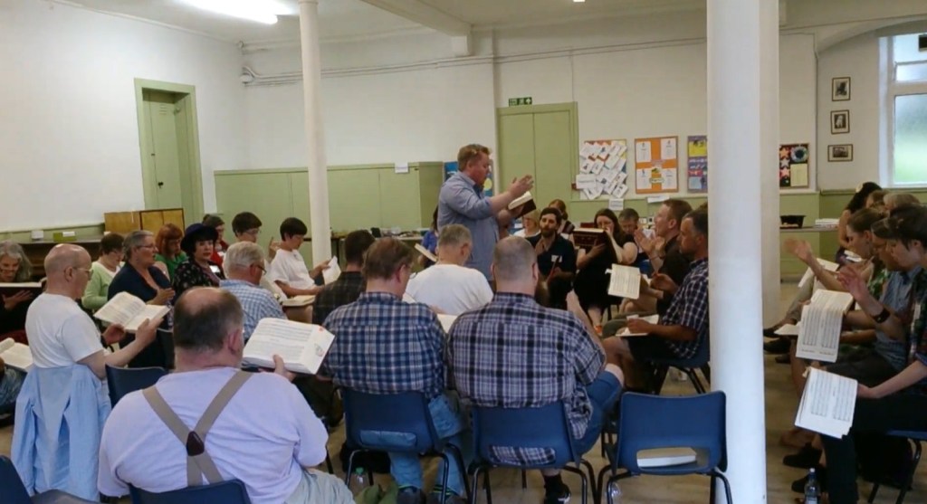 A Sacred Harp square in a large room with white walls and light green cupboards in the background and white pillars supporting the ceiling.