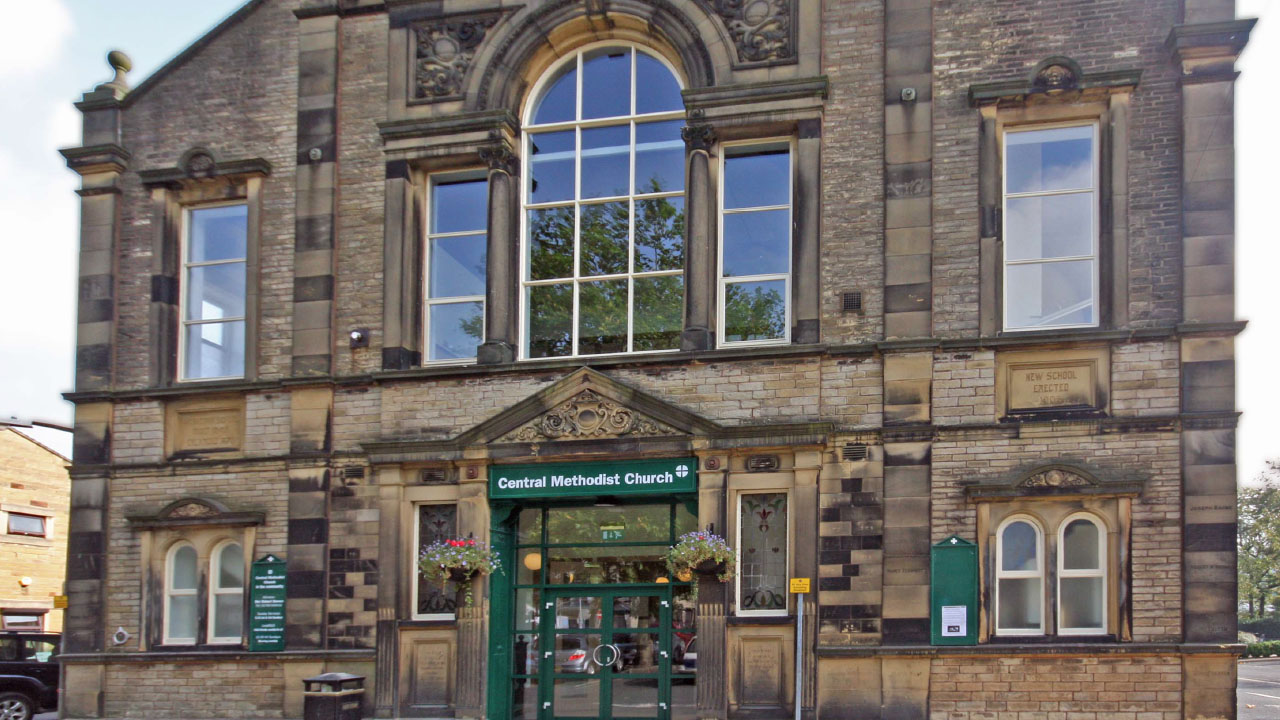 The exterior front face of an imposing stone building. It has a large arched window in the centre on the upper floor, over this, and the double door beneath it, is a panel of carved stone. The sign above the entrance reads 'Central Methodist Church' in white, against the forest green painted background.