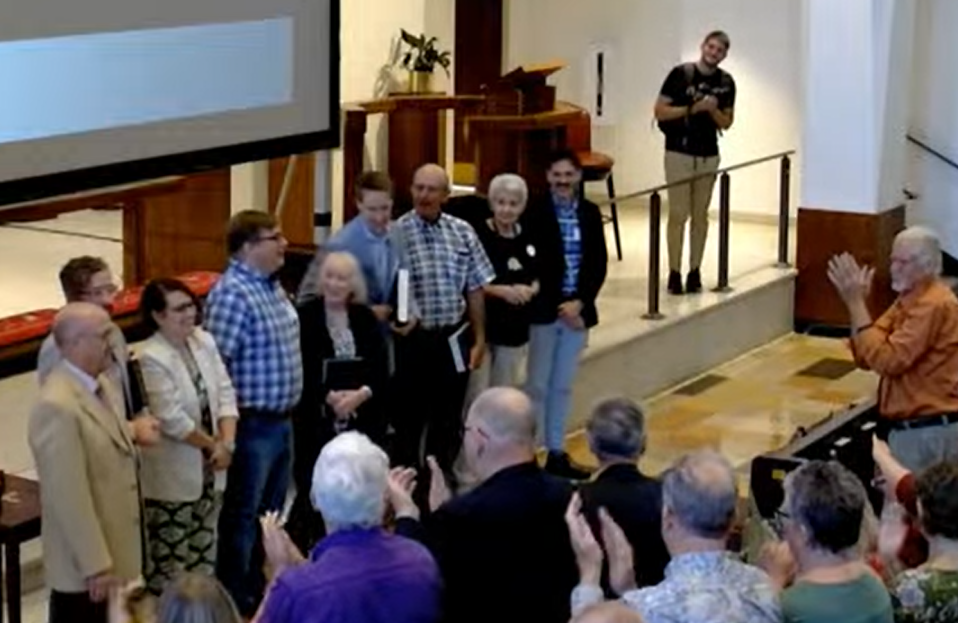 The revision committee for The Sacred Harp 2025 at the Atlanta convention: (left to right) Chair David Ivey, Jesse Karlsberg, Lauren Bock, David Wright, Helen Brown, Richard Ivey, Terry Wootten, Karen Rollins, Nathan Rees.  People in the front rows facing them are applauding enthusiastically!