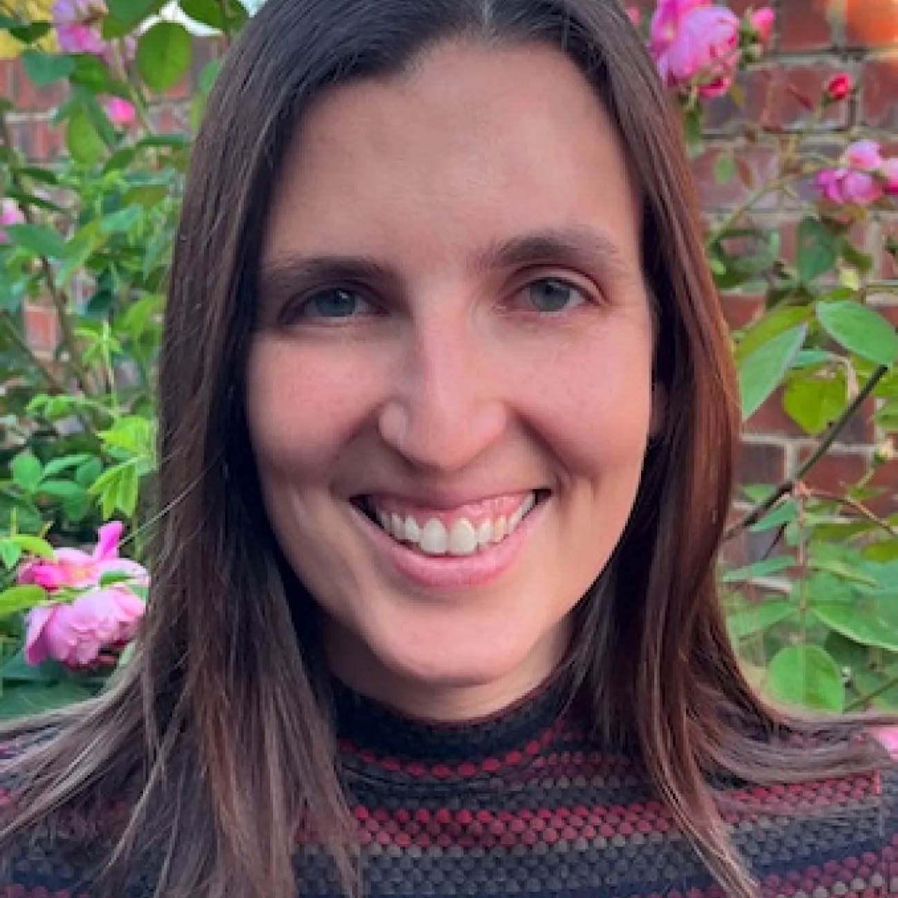 A portrait of Erin Johnson-Williams. Erin is a white woman with dark, straight hair sweeping just below her shoulders and deep blue eyes looking right into the camera. She wears a striped top in deep red and dark grey and a huge beaming smile. 