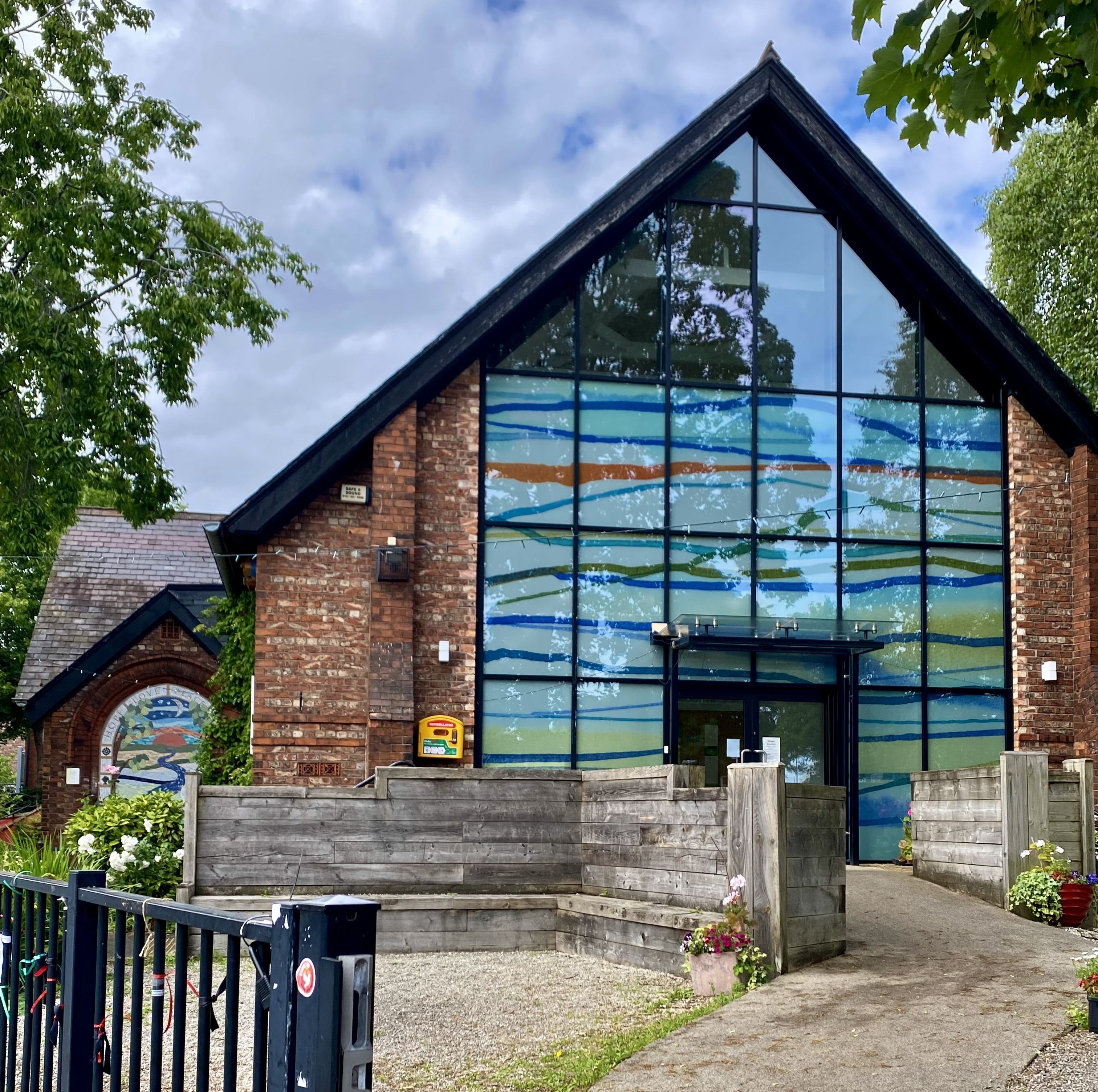 An image of Chorlton Central Church. An inviting-looking red brick building with a huge glass panelled front, made up mostly of contemporary stained glass in white, striped with blue, orange, green and yellow. The sky is blue scattered with fluffy white clouds and where the roof comes to a point we see the tops of large trees surrounding the church. The path ramps up gently towards the double doors. 