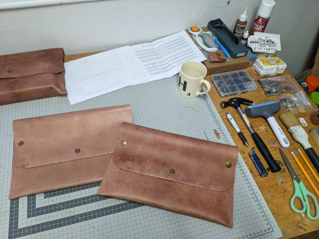 Three reddish-brown booksleeves with gold studs, one already holding a book, on a desk covered with tools, plus a mug and the score of The Cleansing Stream