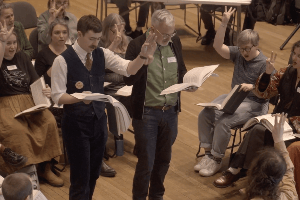 Two men stand in the centre of a square of singers with a large open song book in their hands and their hands raised to indicate they want to sing the third verse. 