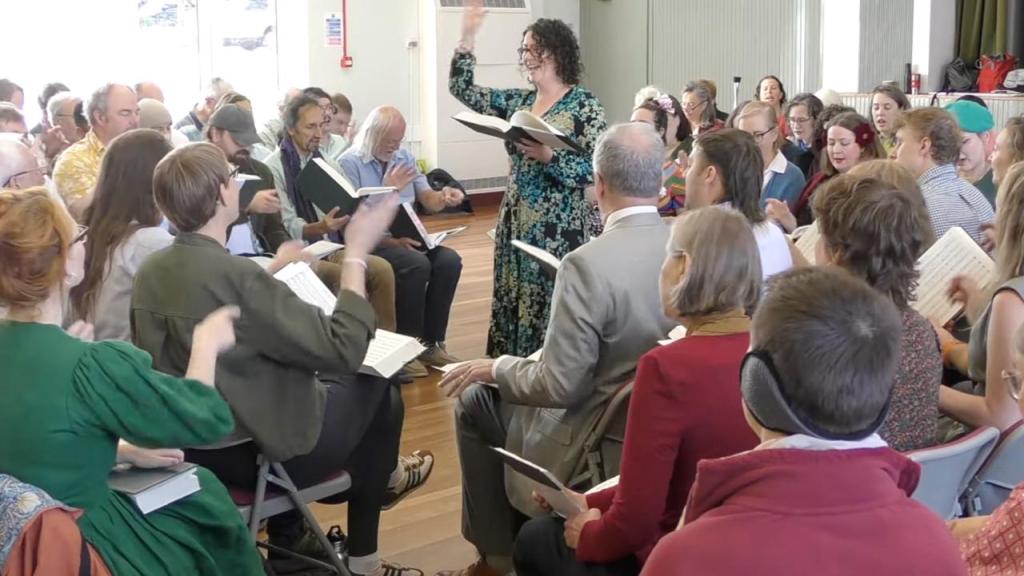 Jo stands in the square in Barley Mow Village Hall, smiling, with her arm raised, as she leads 114 Saint's Delight.