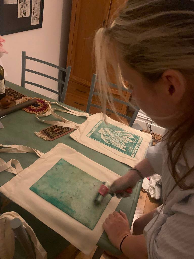 Lucy lino-printing tote bags with a roller. A finished version of the bag, with a green design showing a weary dove, is lying on the table to her right.