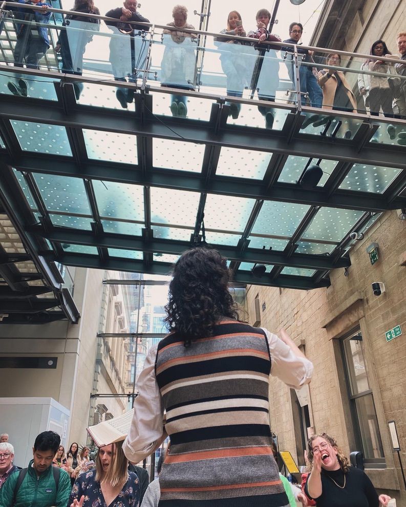 A singer with her back to the camera as she leads a song, with the tenors facing her. Above their heads, visitors to Manchester Art Gallery watch from a glass balcony.