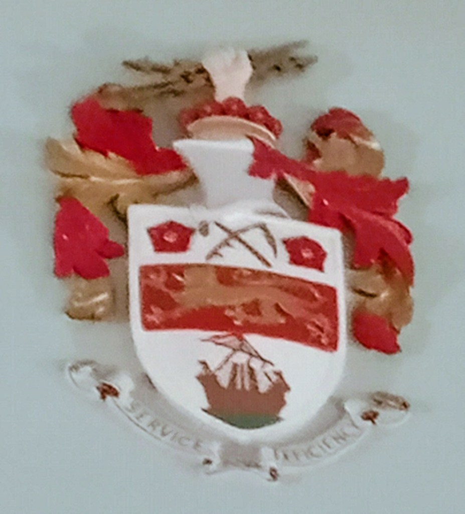 Stretford's coat of arms - on a white background, a flail and scythe between two Lancashire roses, above a golden lion passant on a red stripe, above a galley ship. The motto below reads SERVICE EFFICIENCY