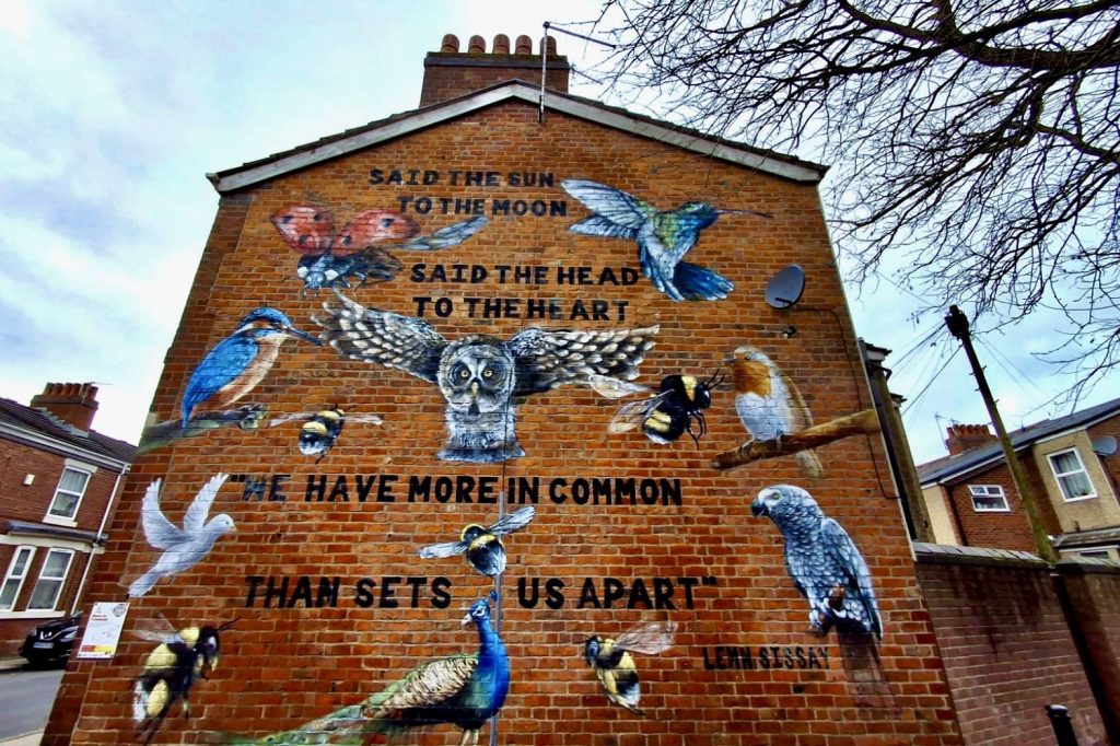 The end wall of a red brick house on which a huge mural is painted. Painted nature details - an owl, a bee, a kingfisher, a dove -intersperse a line of poetry by Lemn Sissay which reads "Said the sun to the moon, said the head to the heart, we have more in common than sets us apart".