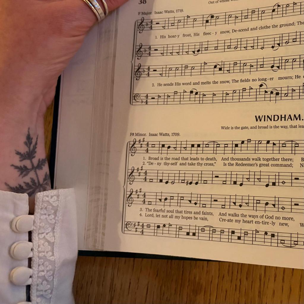 A page of the Sacred Harp 2025 edition, with assorted black and white blobs. To the right, an arm with a delicate leaf tattoo emerging from under a white sleeve.