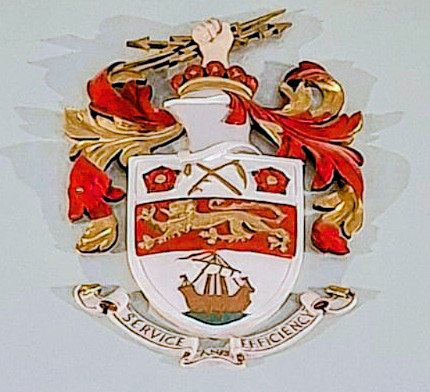 Stretford's coat of arms - on a white background, a flail and scythe between two Lancashire roses, above a golden lion passant on a red stripe, above a galley ship. The motto below reads SERVICE AND EFFICIENCY. Photo by Susanna Cox